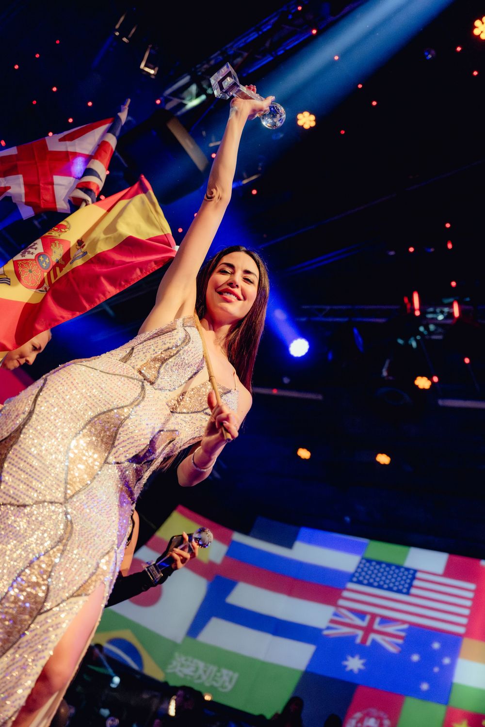 A performer in a sparkly dress shines on stage with international flags in the background.