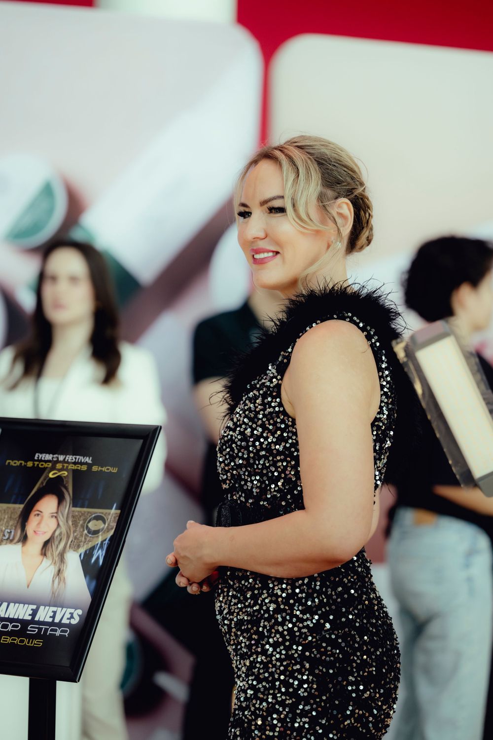 A woman in a sparkly black evening gown smiles while holding a promotional sign.
