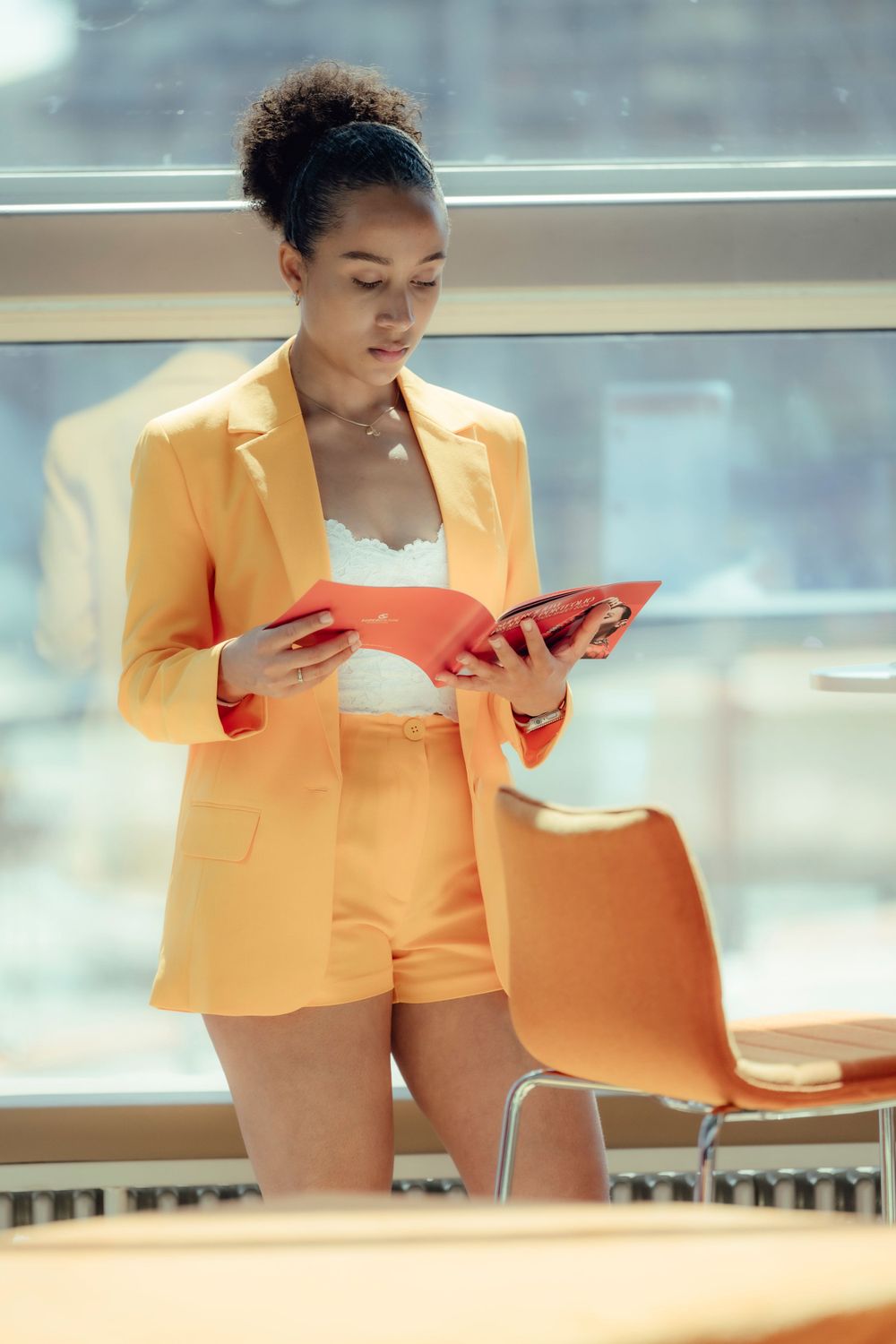 A woman in a bright orange blazer and matching shorts reads documents in an office setting.