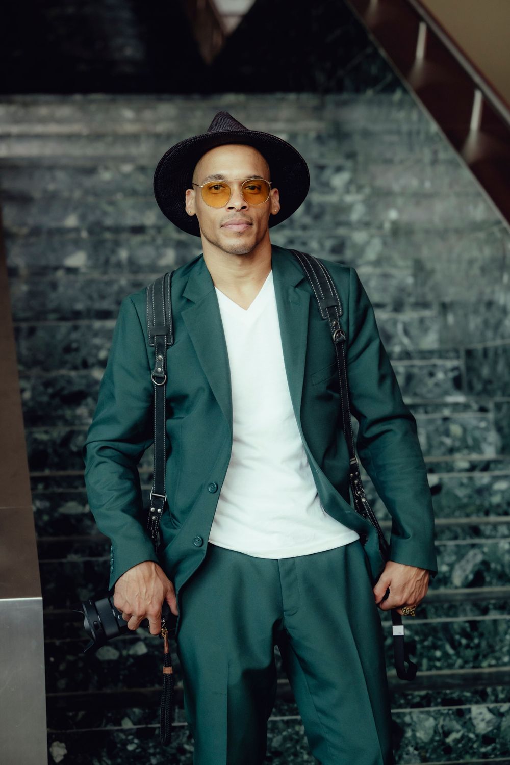 A stylish man in a green suit and hat poses against stone steps.
