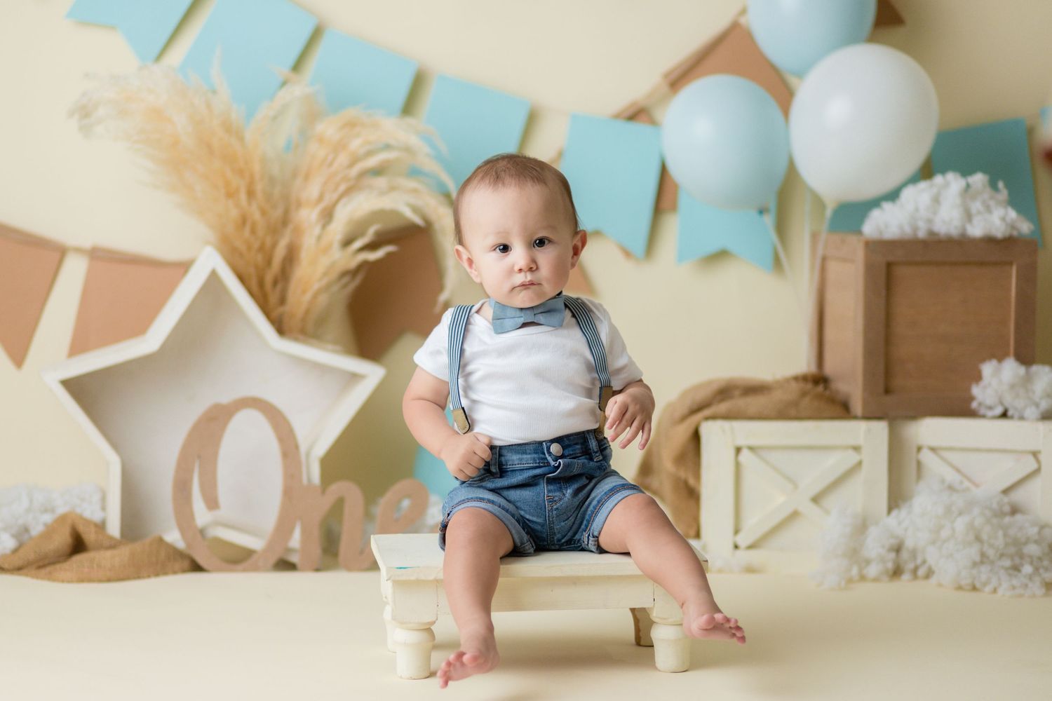 Baby in blue shorts sits on chair with blue balloons and rustic decorations in background.