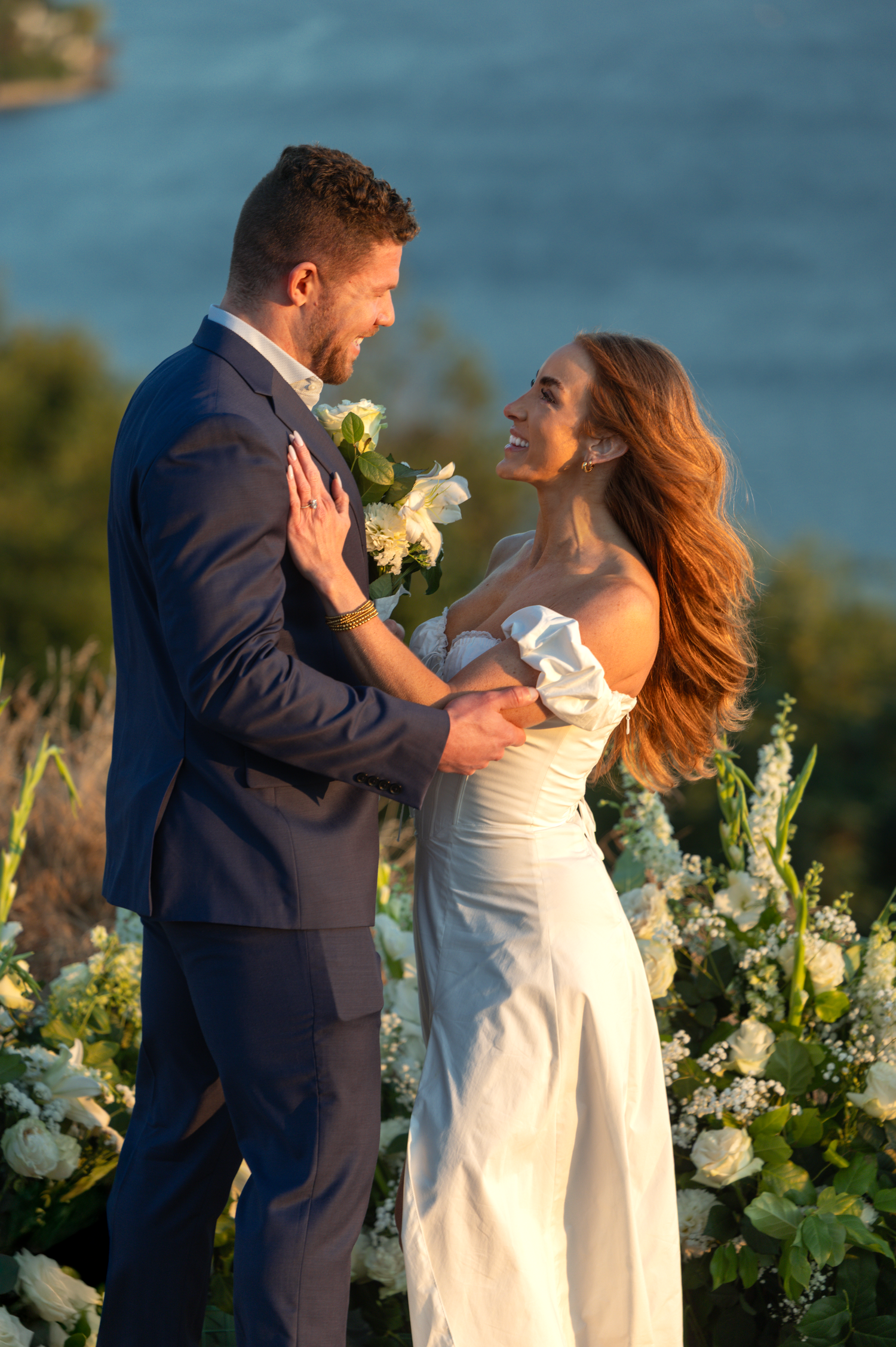 A romantic outdoor wedding moment at sunset by the ocean with white floral decorations and stunning natural light.