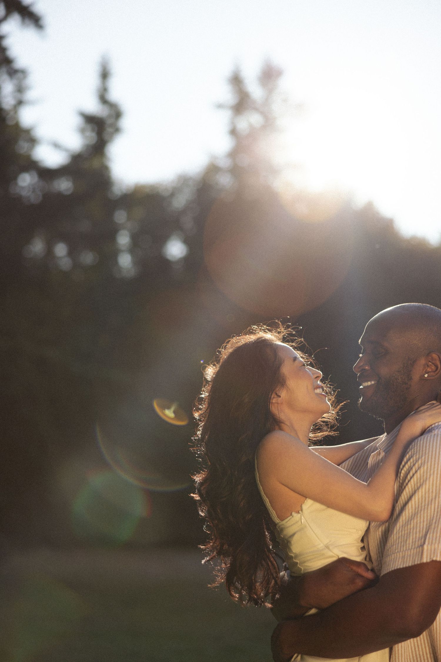 A romantic couple embraces outdoors in warm sunset light with lens flare glowing through trees.