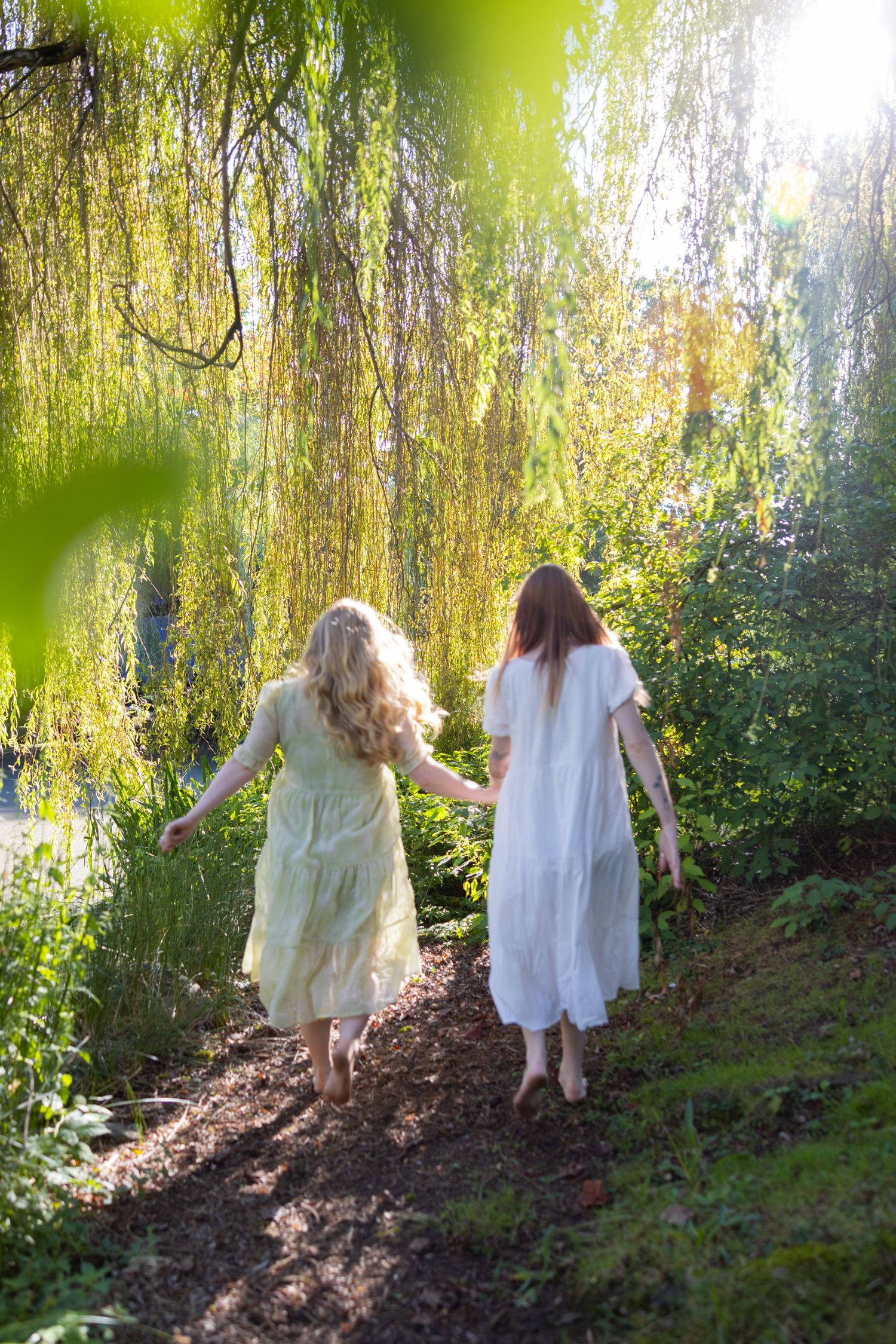 Two figures in white dresses walk barefoot along a sunlit garden path beneath hanging willow branches.
