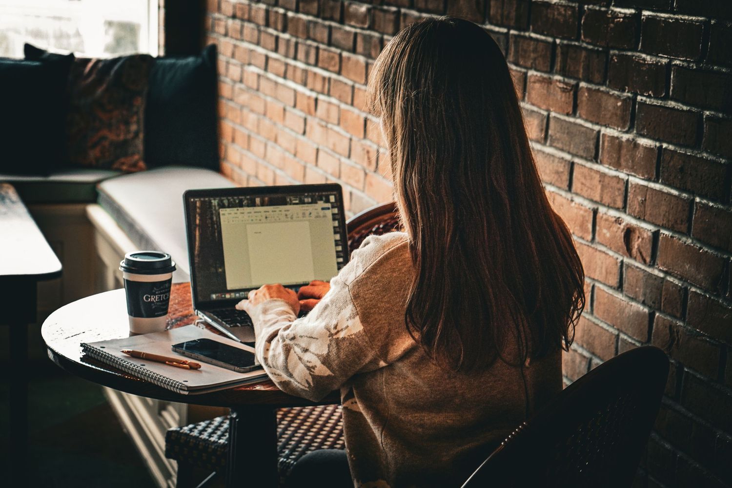 Person working on laptop in coffee shop with exposed brick wall and takeout cup.