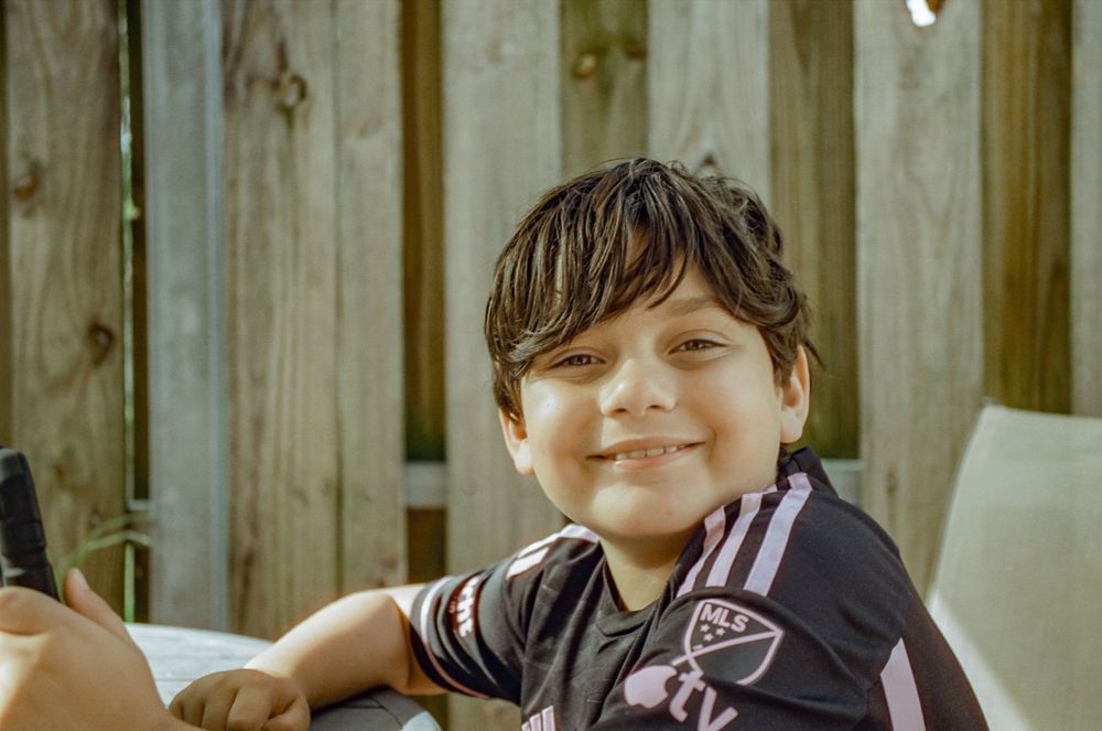Young person wearing a black jersey smiling warmly outdoors against a wooden fence background.