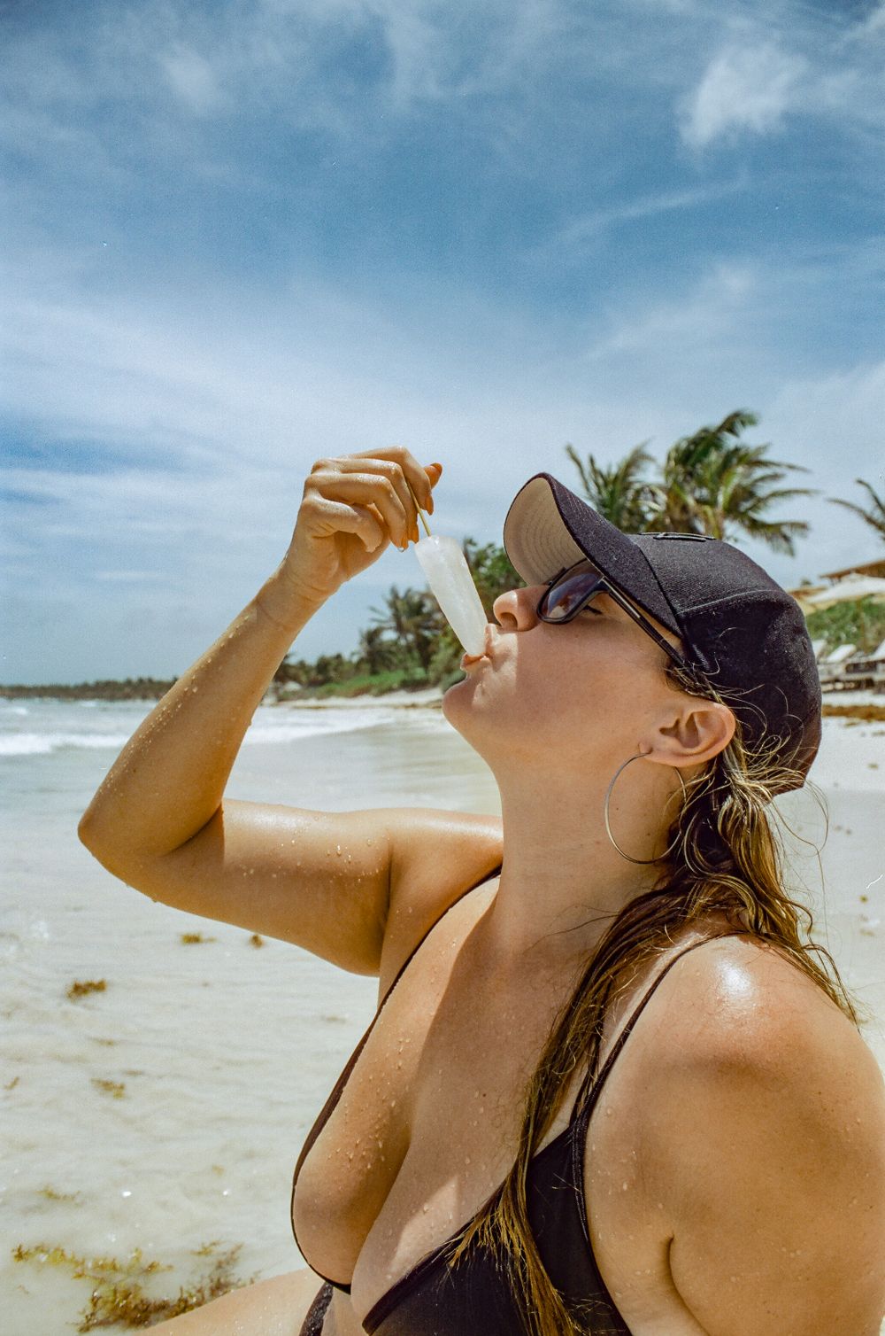A beachgoer in a baseball cap enjoys a refreshing drink against a tropical beach backdrop.