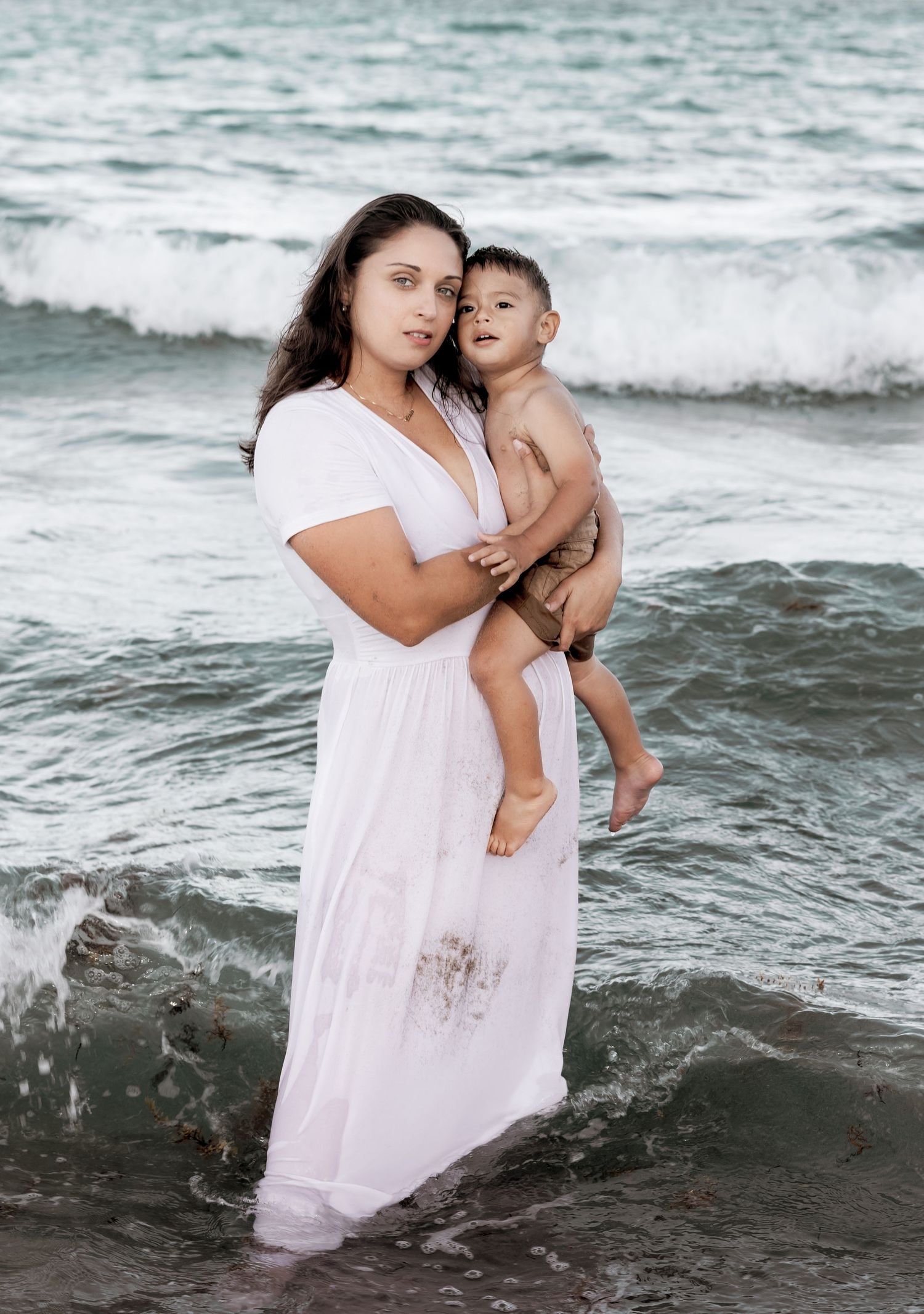 Woman in white dress holding child while standing in ocean waves.
