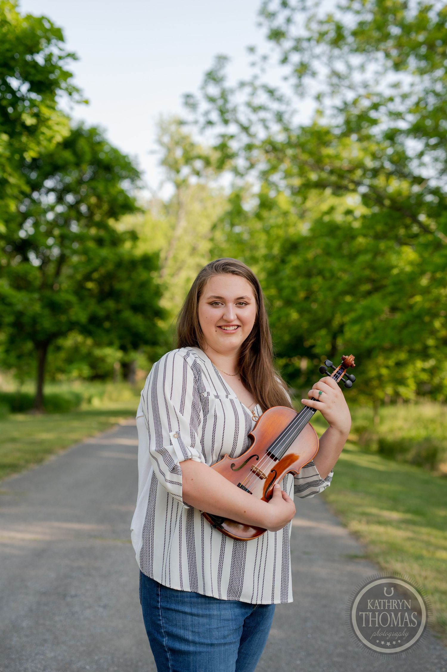 Violinist in casual attire poses with instrument on sunny outdoor path.