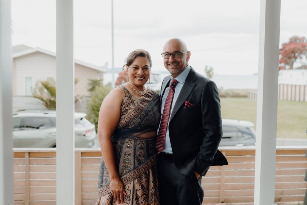 A couple poses for a photo on an outdoor porch, the woman wearing a patterned dress and the man in a dark suit.