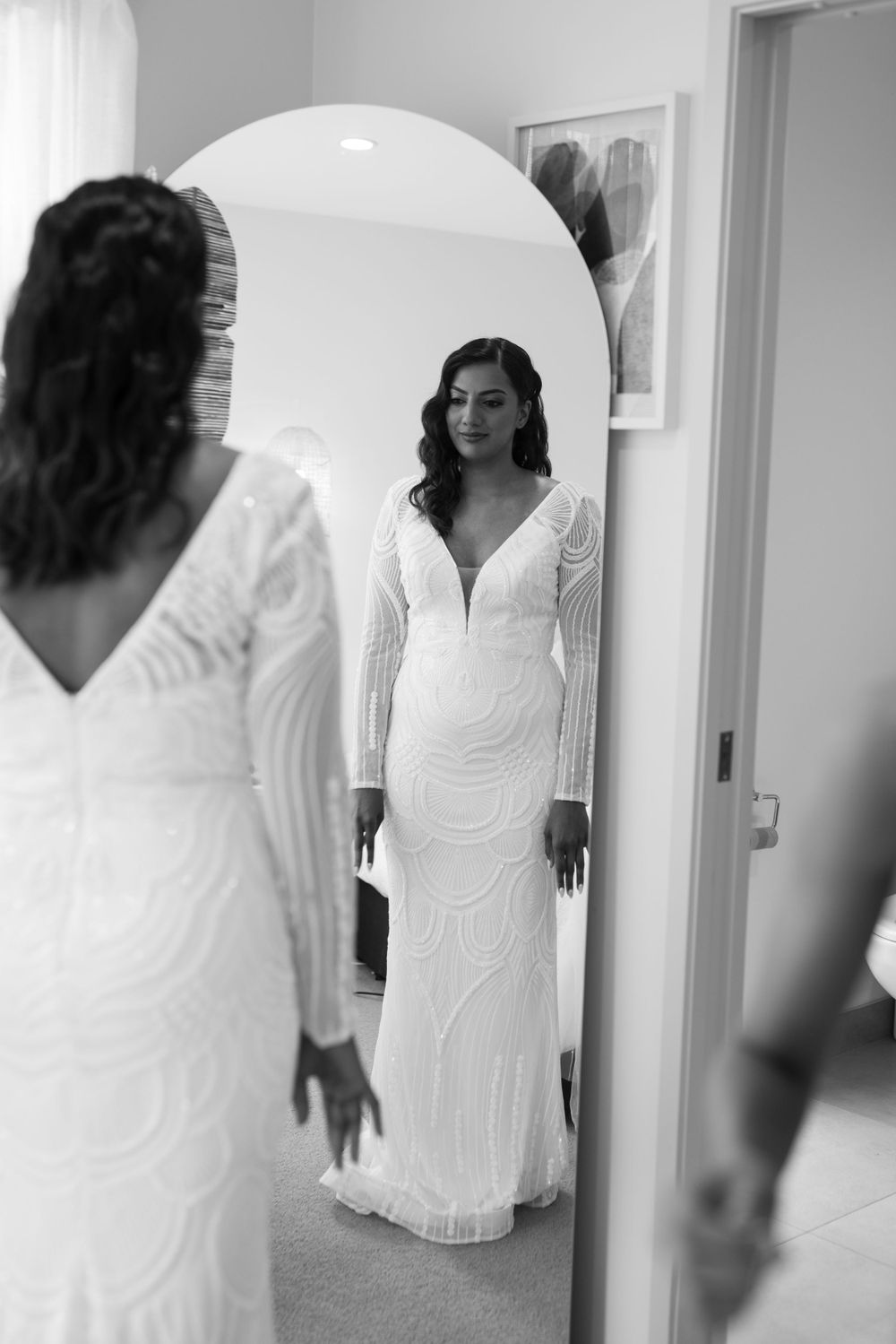 A bridal gown fitting session reflected in an oval mirror showing intricate lace details.