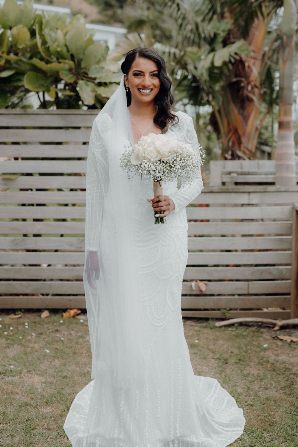A bride in a long-sleeved white gown holds a baby's breath bouquet against a wooden fence backdrop.