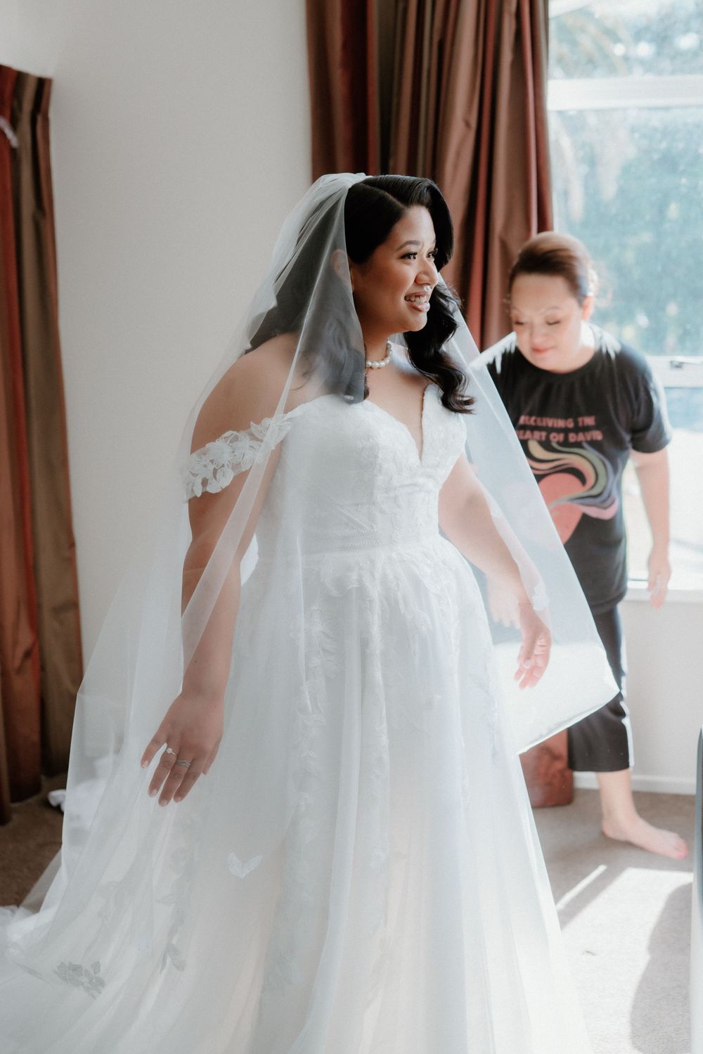 A bride in a white off-shoulder wedding dress stands in a room with natural lighting.