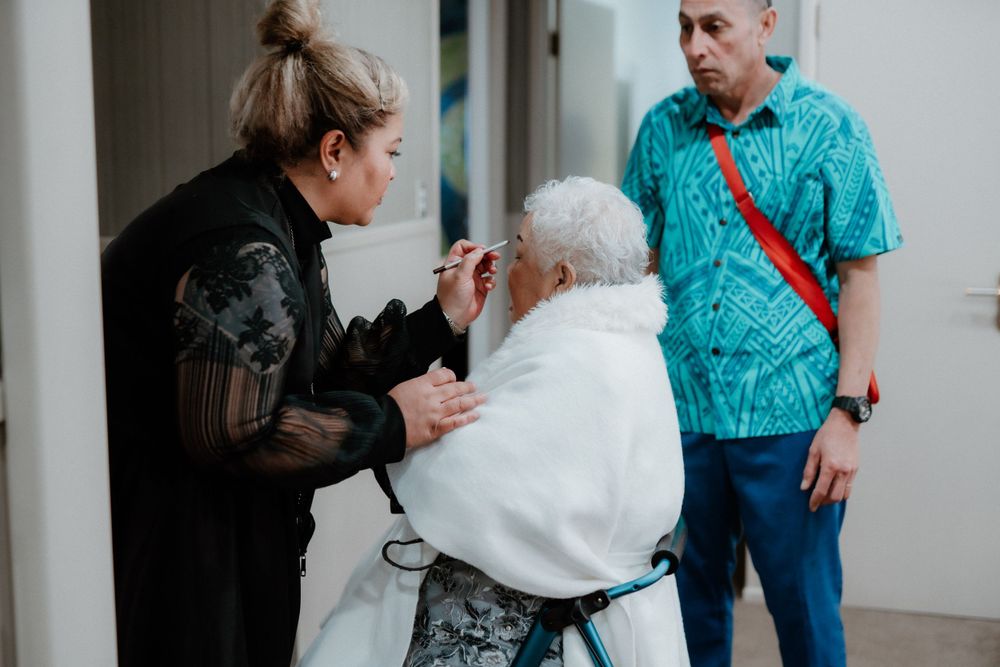 A stylist applies makeup to a person in a white robe while another looks on.
