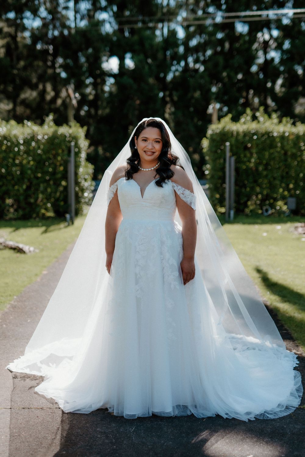 A bride in an elegant white A-line wedding gown and long veil poses outdoors on a garden path.