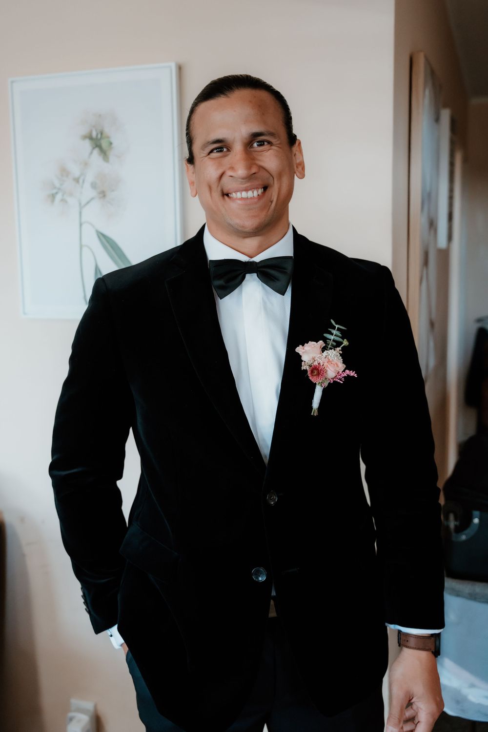 A dapper groom in a classic black tuxedo and bow tie with a pink boutonniere smiles at the camera.