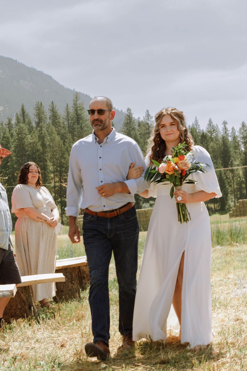 Bride in white dress with summer bouqet and her father walking down the aisle for wedding ceremony, family member smiling in the background.