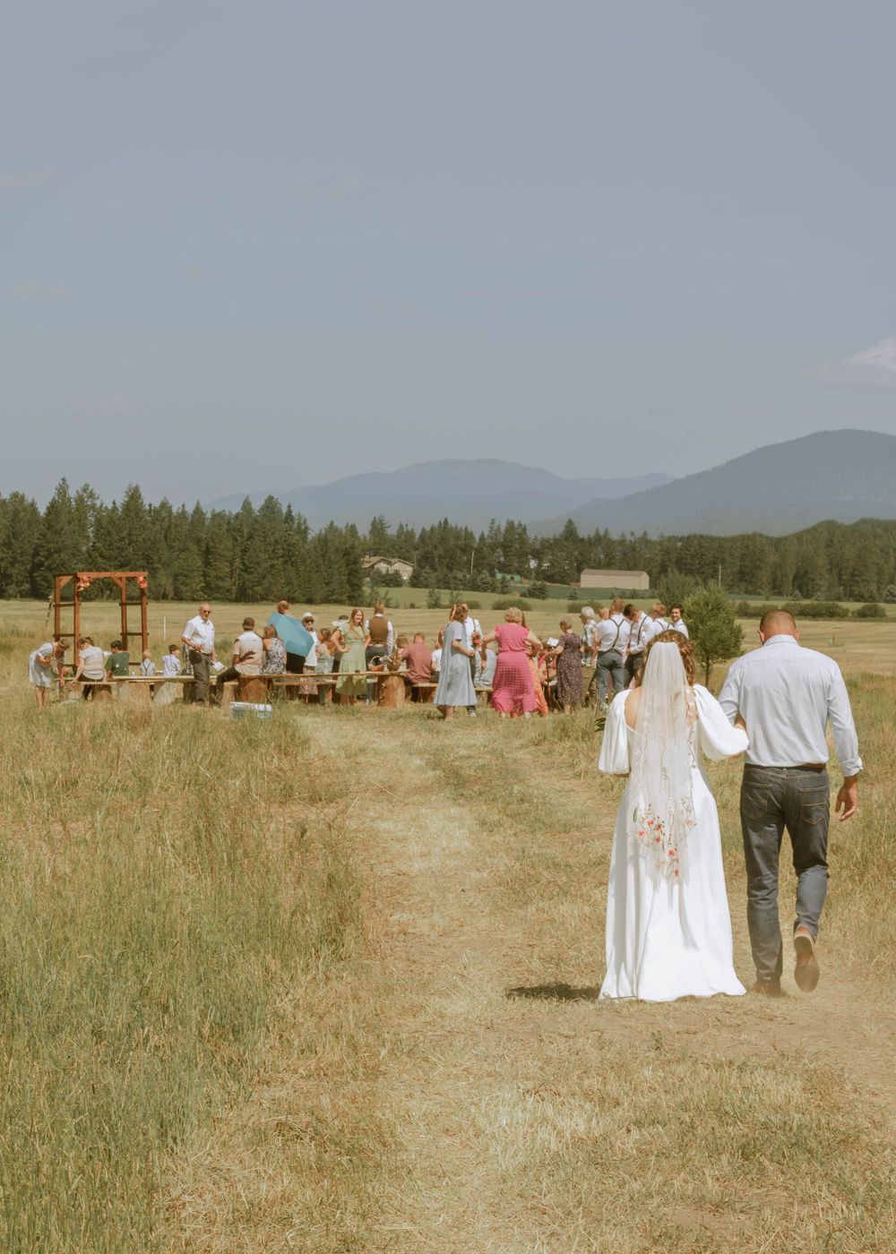 View of bride and her father walking down grassy aisle towards groom and ceremony location, mountains and trees in background.