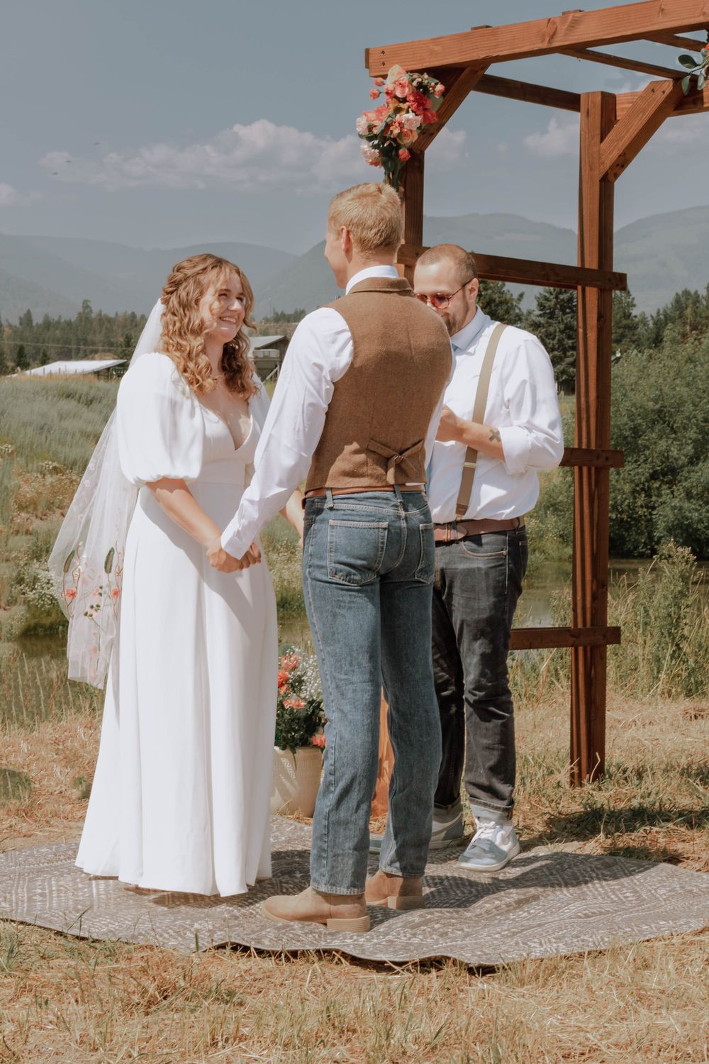 Bride and groom at altar with officiant, groom's brother, grassy mountain scenery.