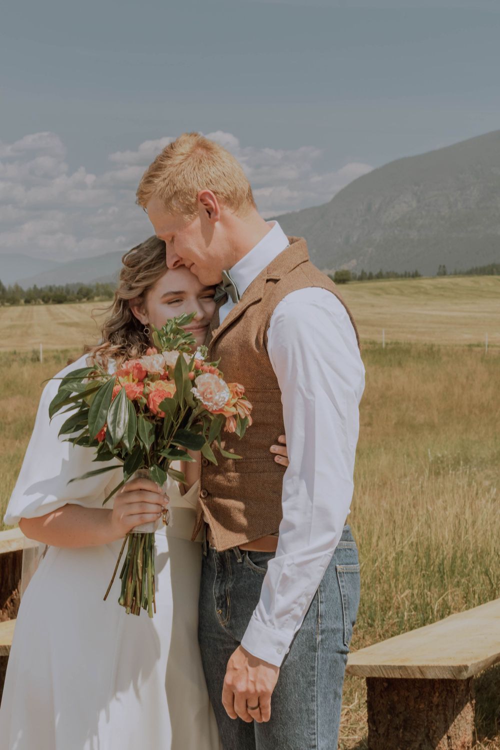 Newly married couple embracing after wedding ceremony with plains, mountains and tree line in view beyond.