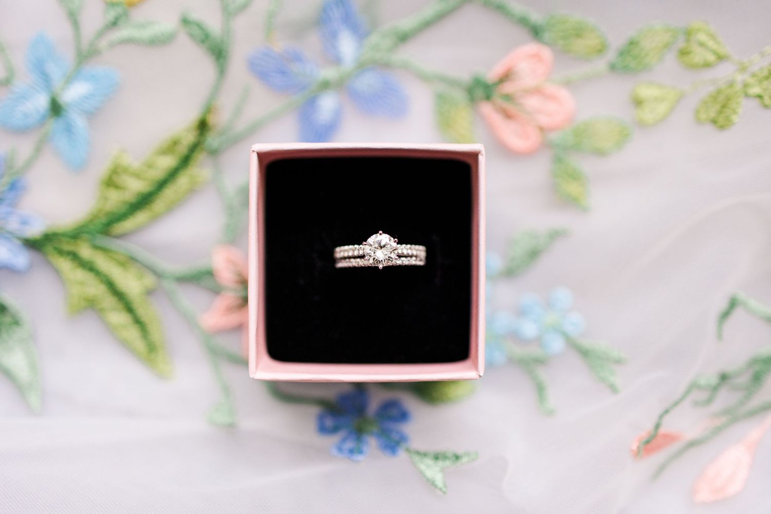 An elegant engagement ring displayed in a black box against a floral patterned background.