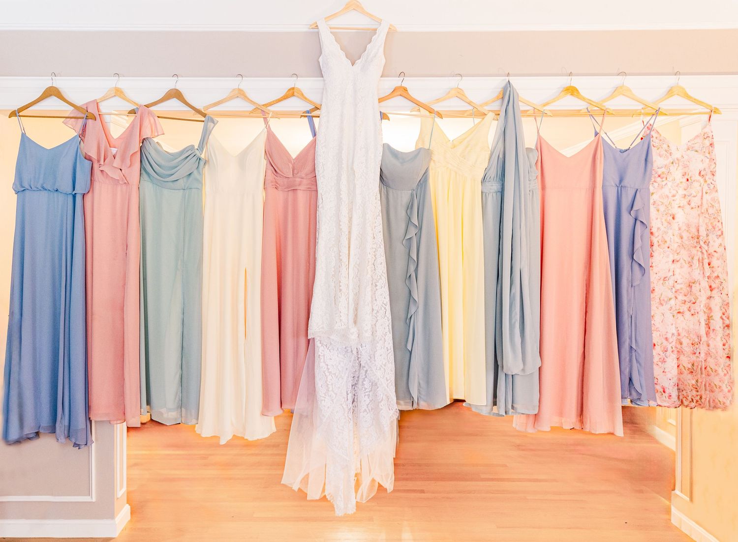 Colorful bridesmaid dresses in pastel shades hanging on a rack with white wedding gown in center.