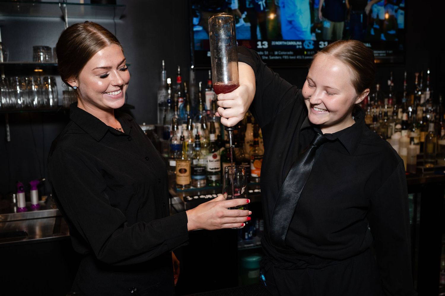 Two bartenders in black attire pour drinks behind a bar counter in a dimly lit establishment.