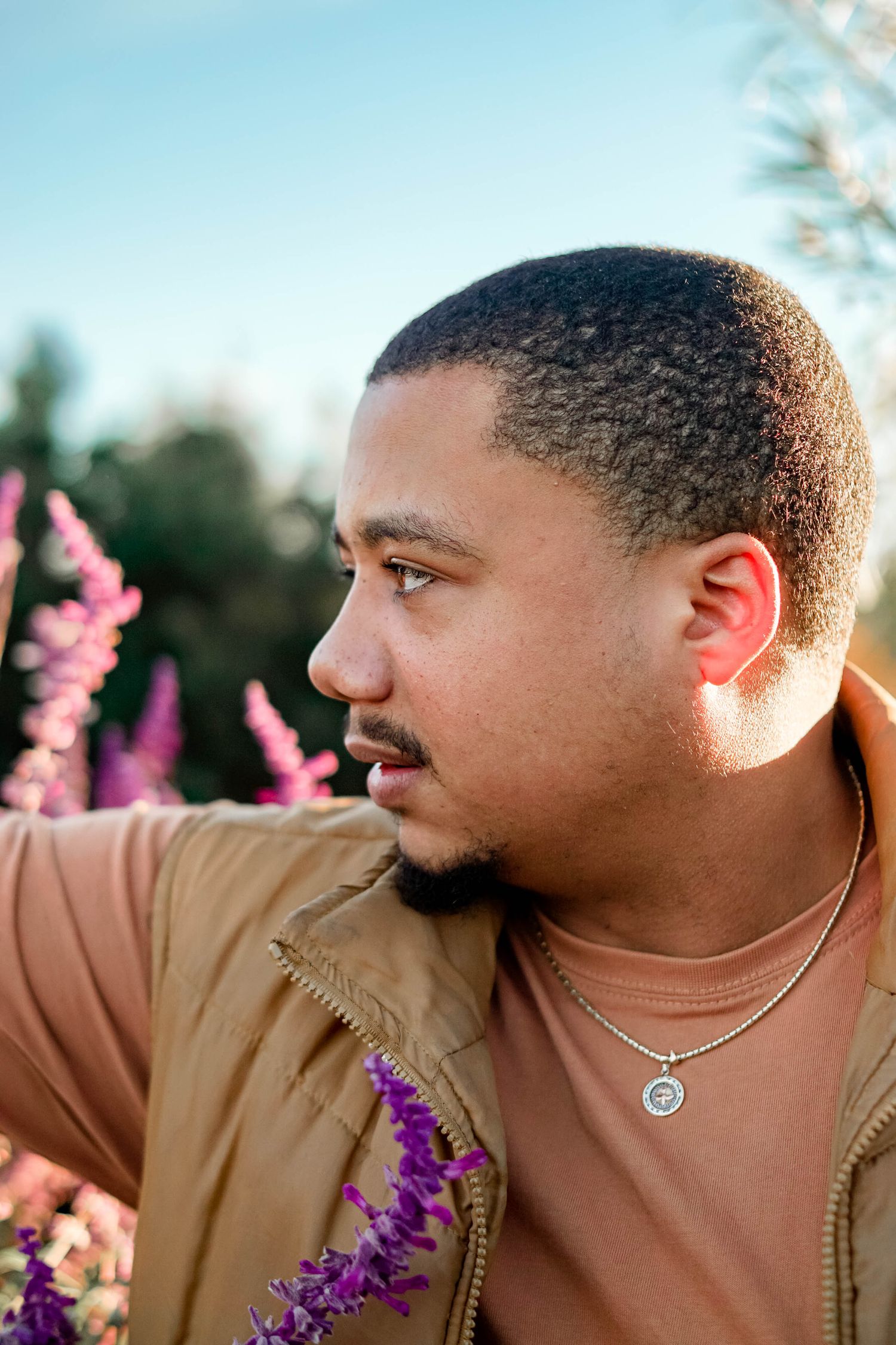 A young man with a necklace gazes sideways surrounded by purple wildflowers in warm sunlight.
