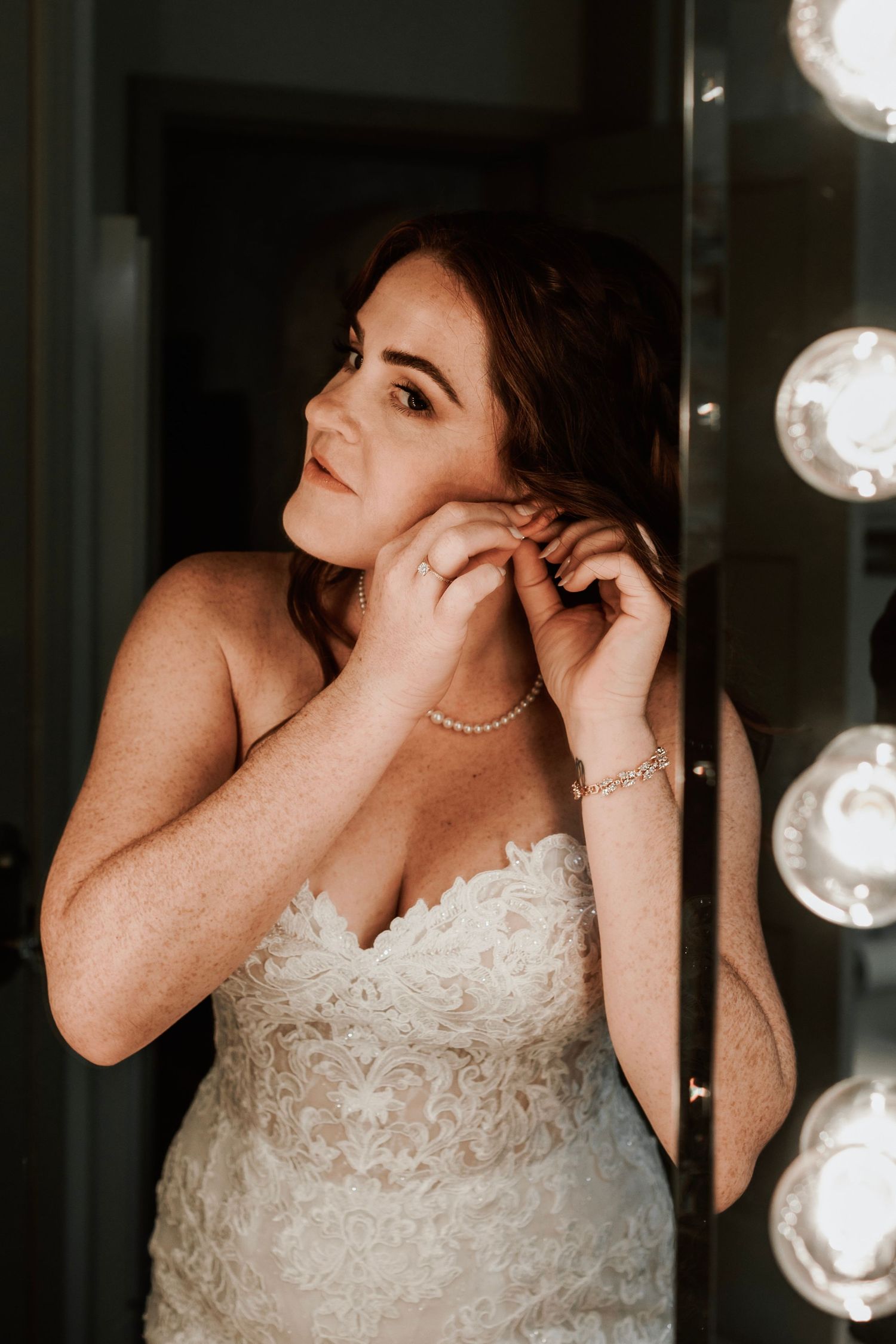 Bride in strapless lace gown putting on earrings in front of a Hollywood vanity mirror.