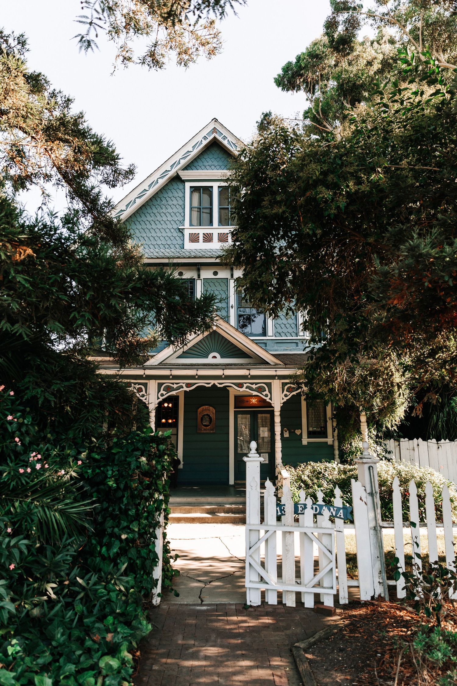 Charming Victorian-style home with teal siding and white picket gate surrounded by lush trees.
