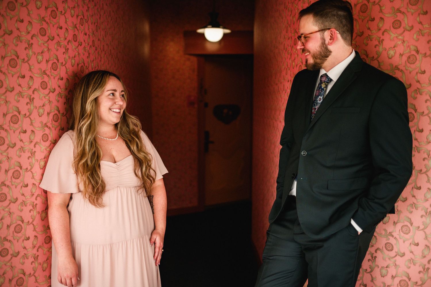 Bride in blush pink dress and groom in dark suit share a smile in a vintage wallpapered hallway.