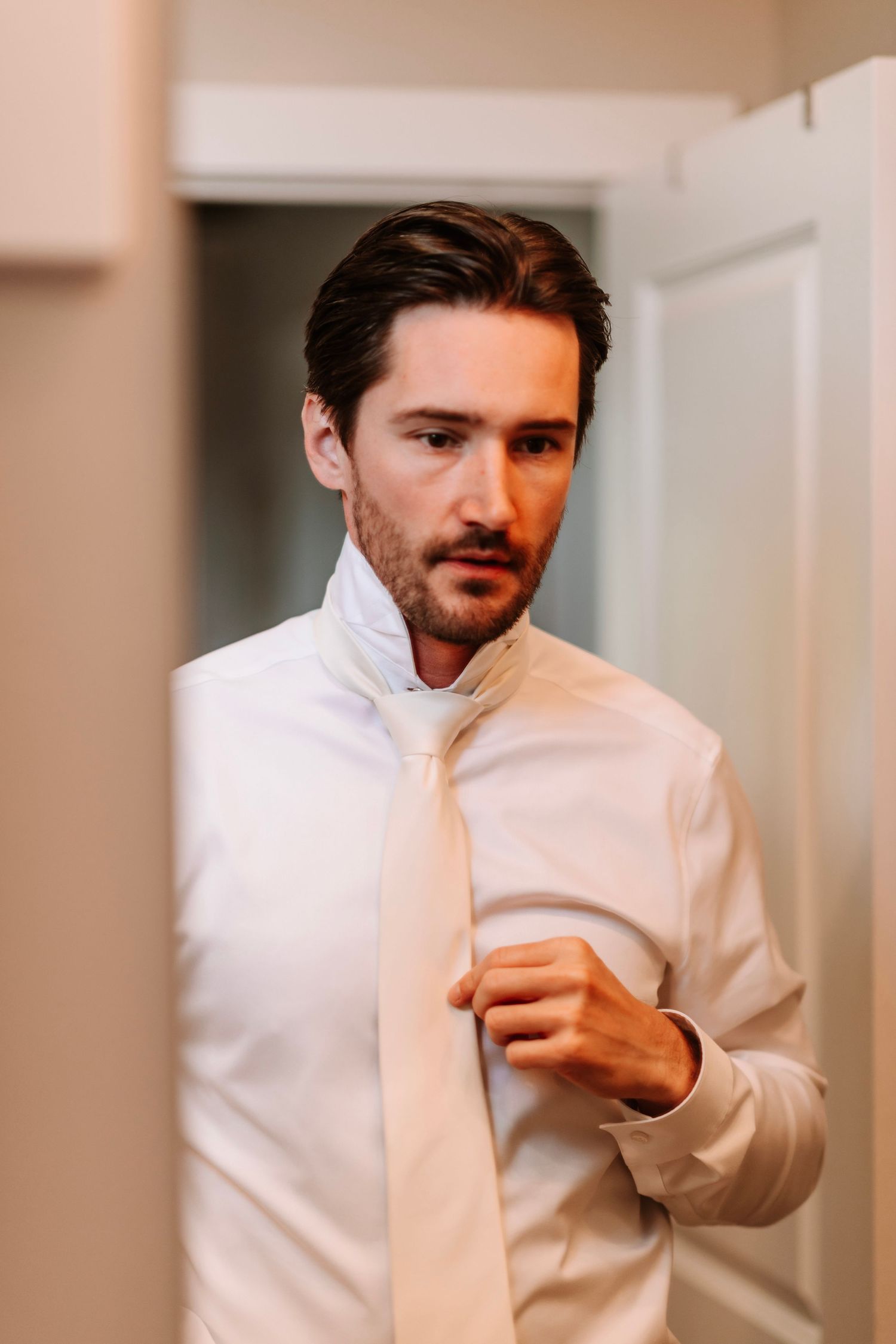 Handsome groom in white dress shirt adjusting his tie while getting ready in front of a mirror.