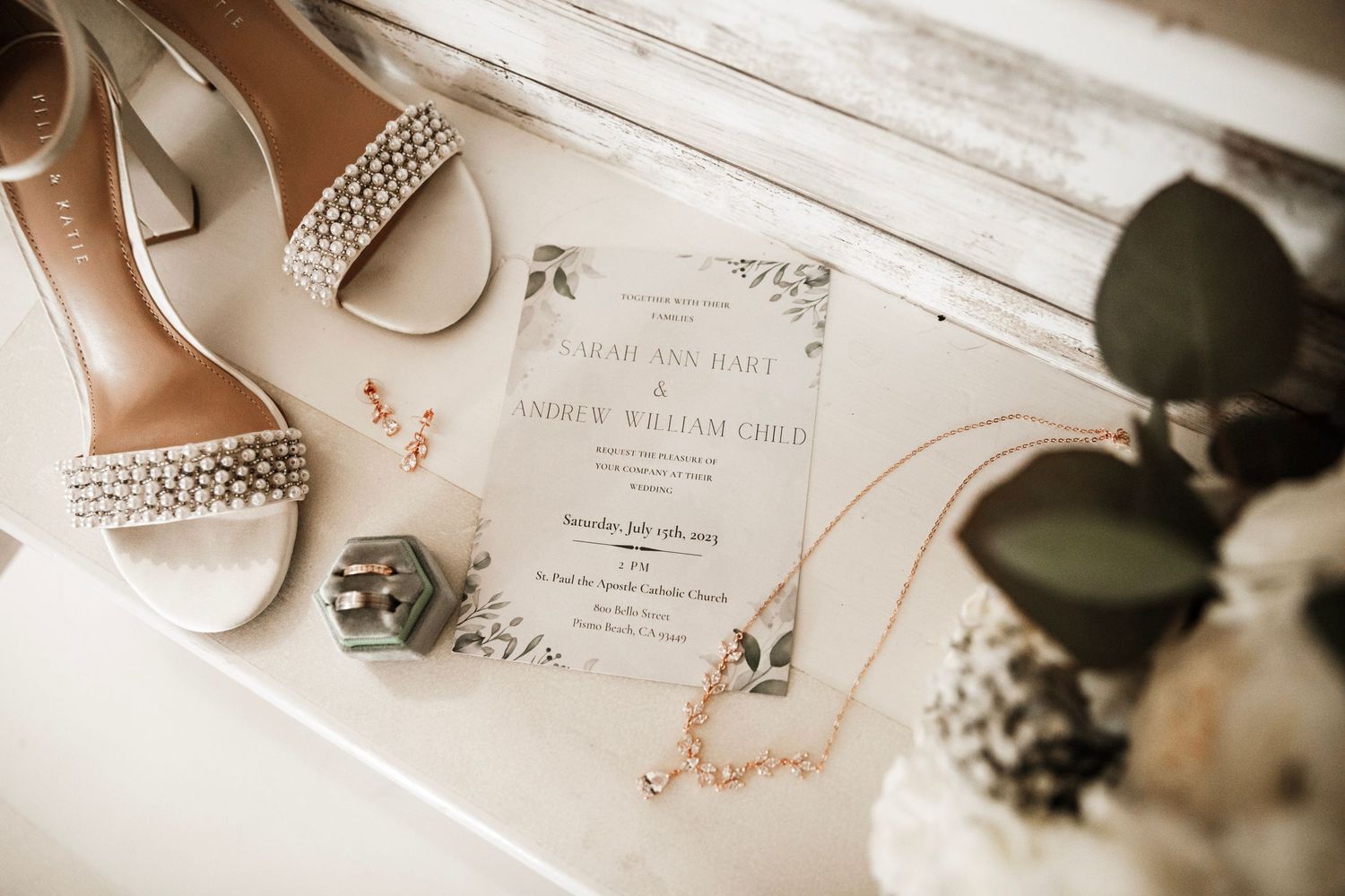 Elegant wedding flat lay featuring embellished heels, gold jewelry, green ring box, and floral invitation suite.