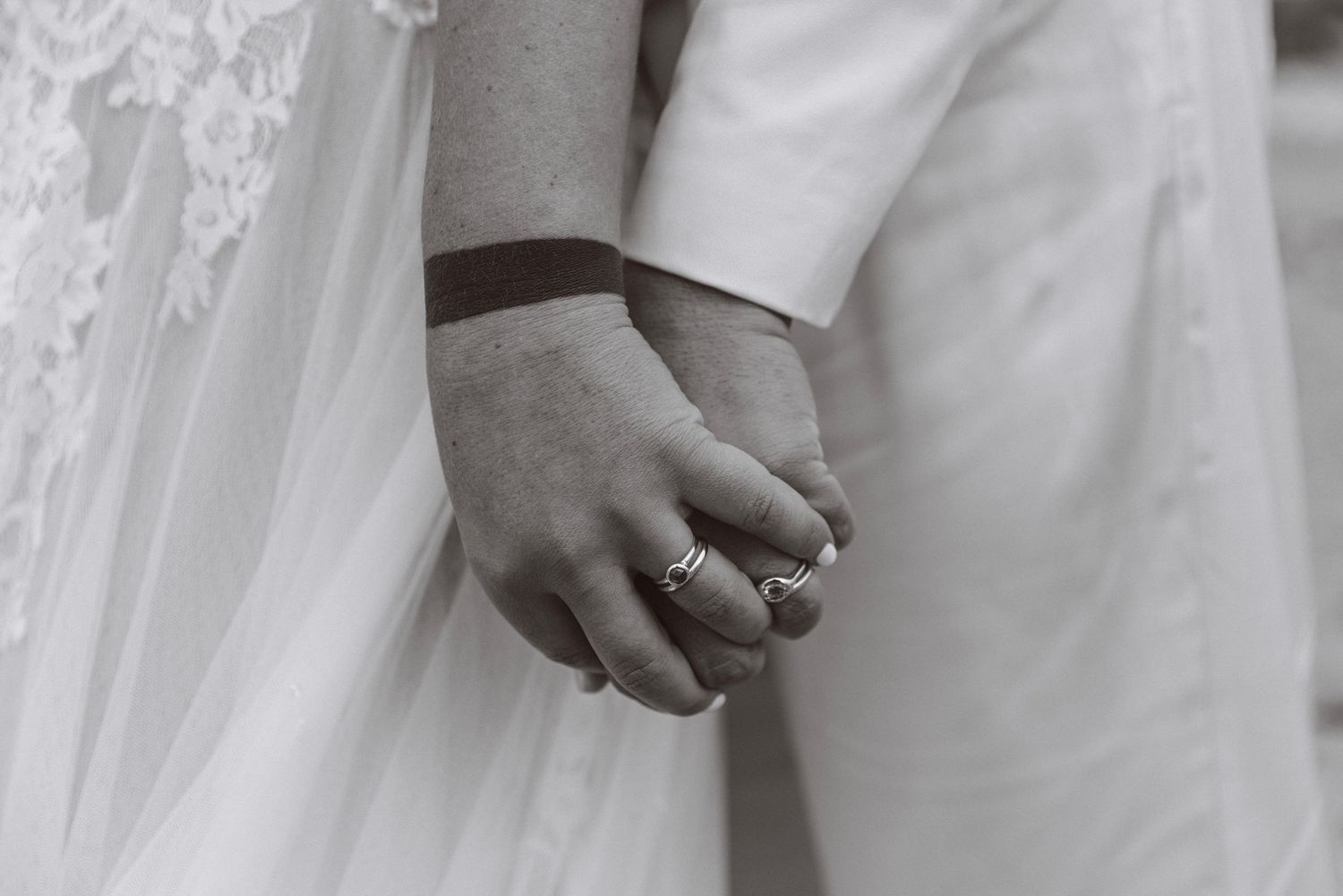 Black and white close-up of bride and groom holding hands showing wedding rings and bracelets.