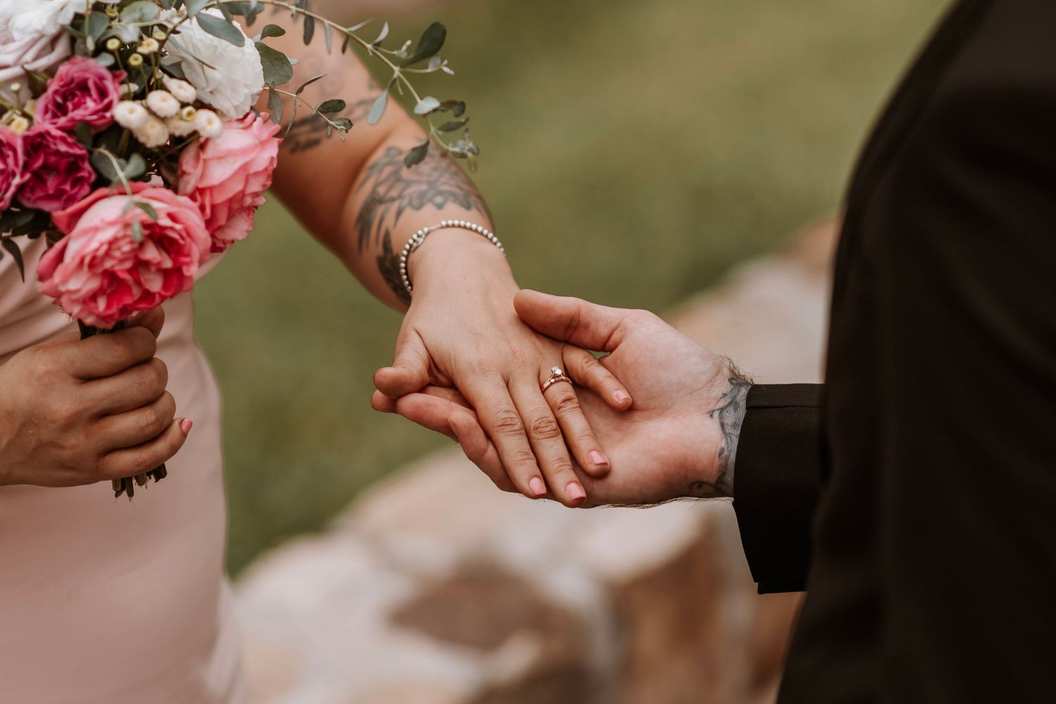 Close-up of a couple holding hands, showcasing a wedding ring and floral tattoo with a pink bouquet.