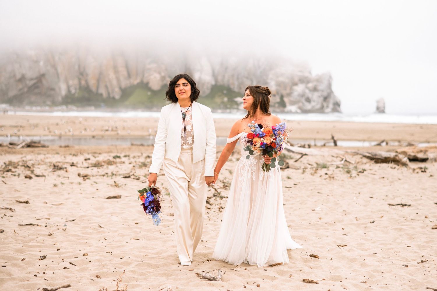 Two brides holding colorful bouquets walk hand in hand on sandy beach with misty sea stacks backdrop.