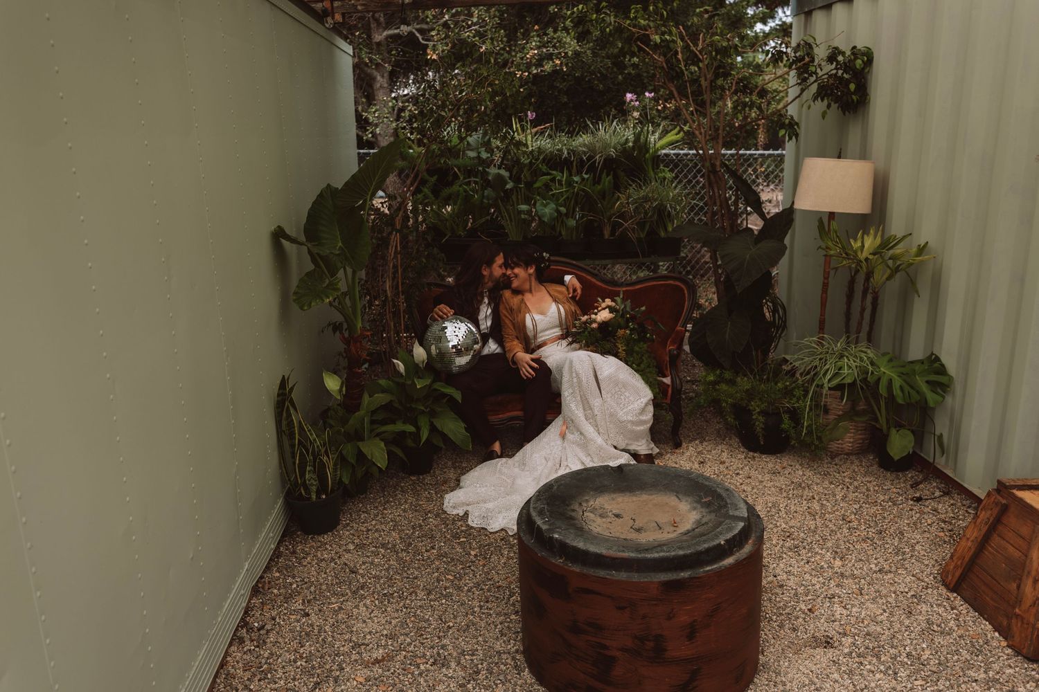 Bride in white gown sits among lush tropical plants in a cozy outdoor garden courtyard setting.