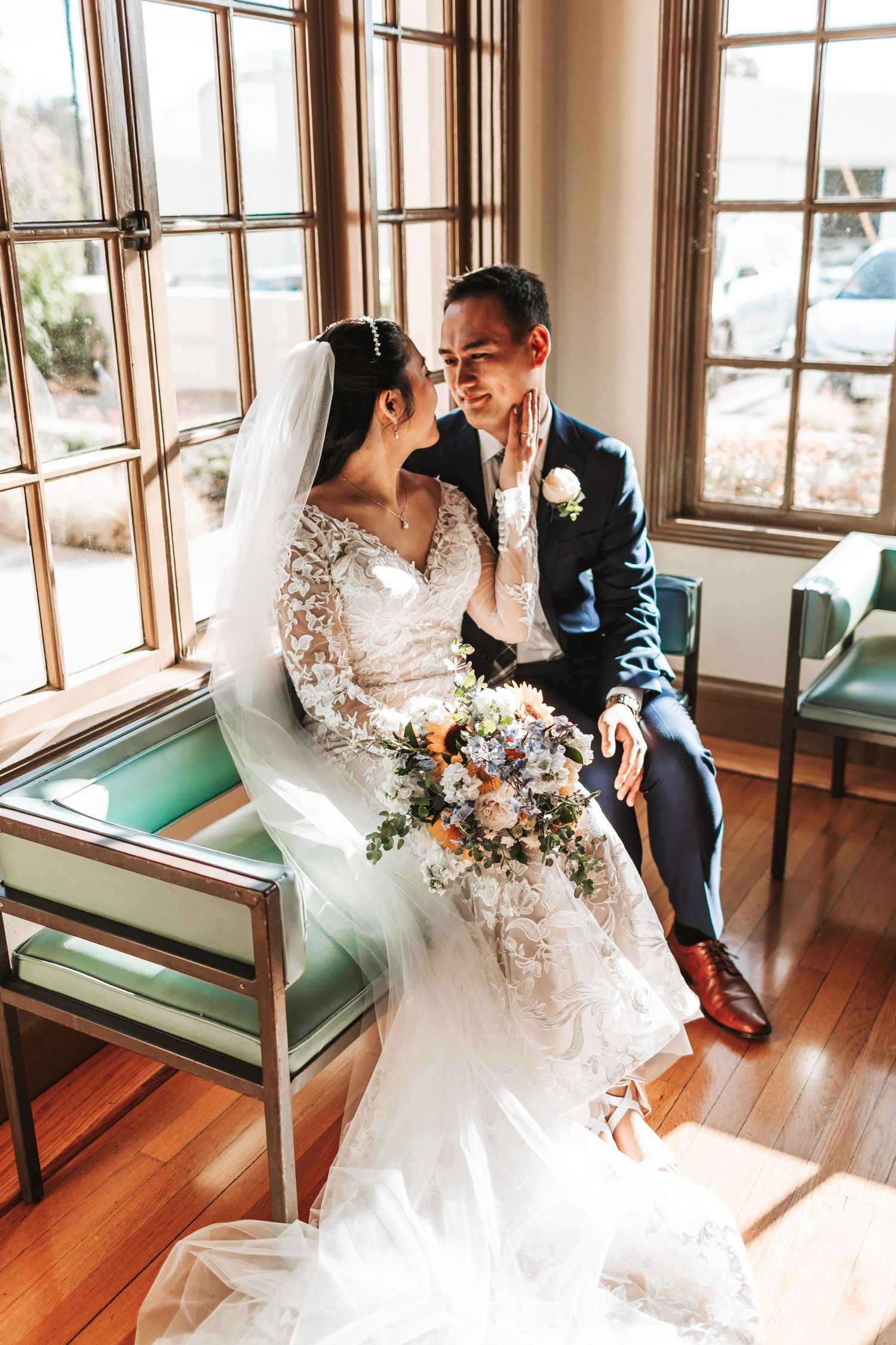 Bride and groom sit intimately by sunlit window with colorful bridal bouquet indoors.
