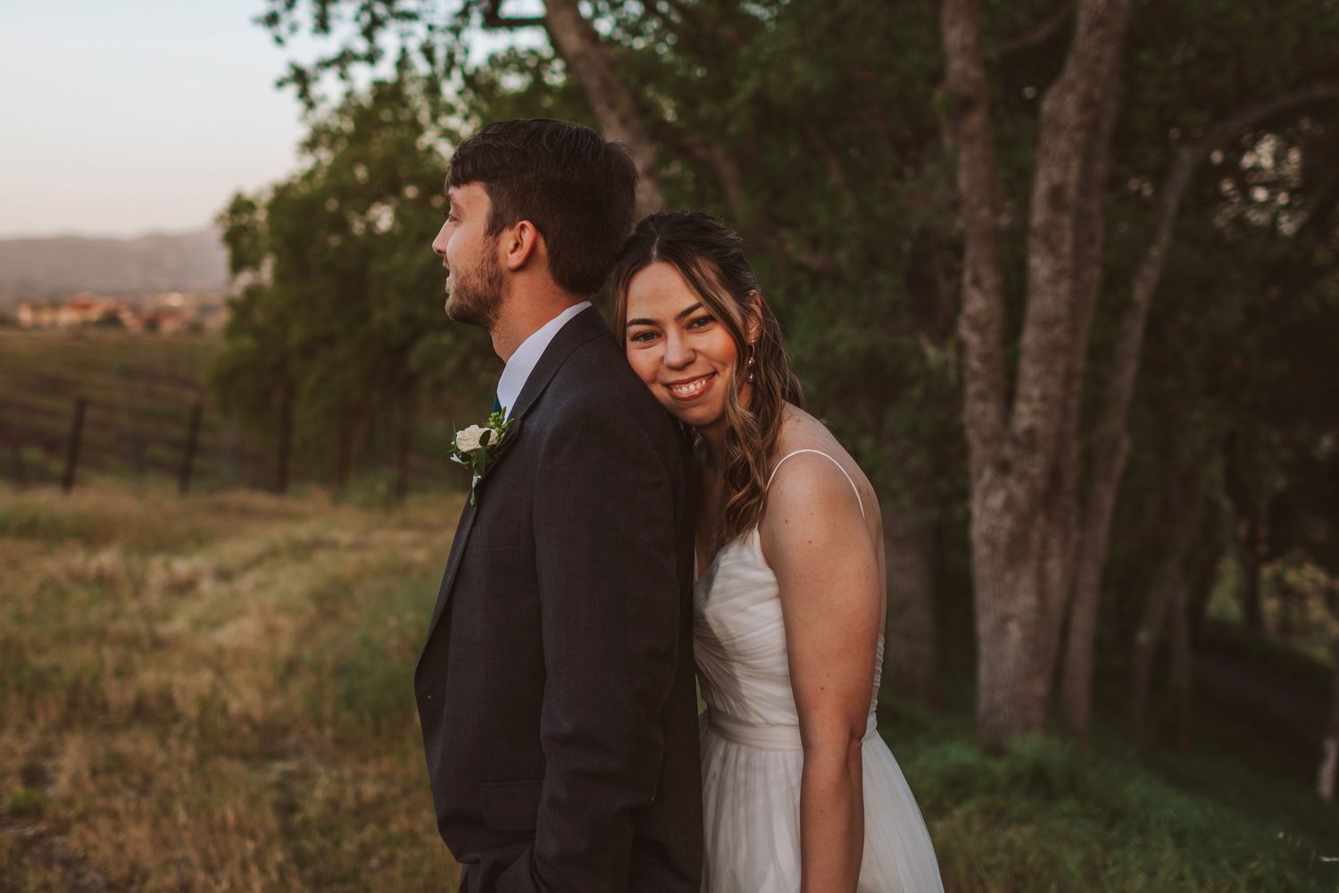 Smiling bride in white gown stands back-to-back with groom in a golden sunset vineyard field.