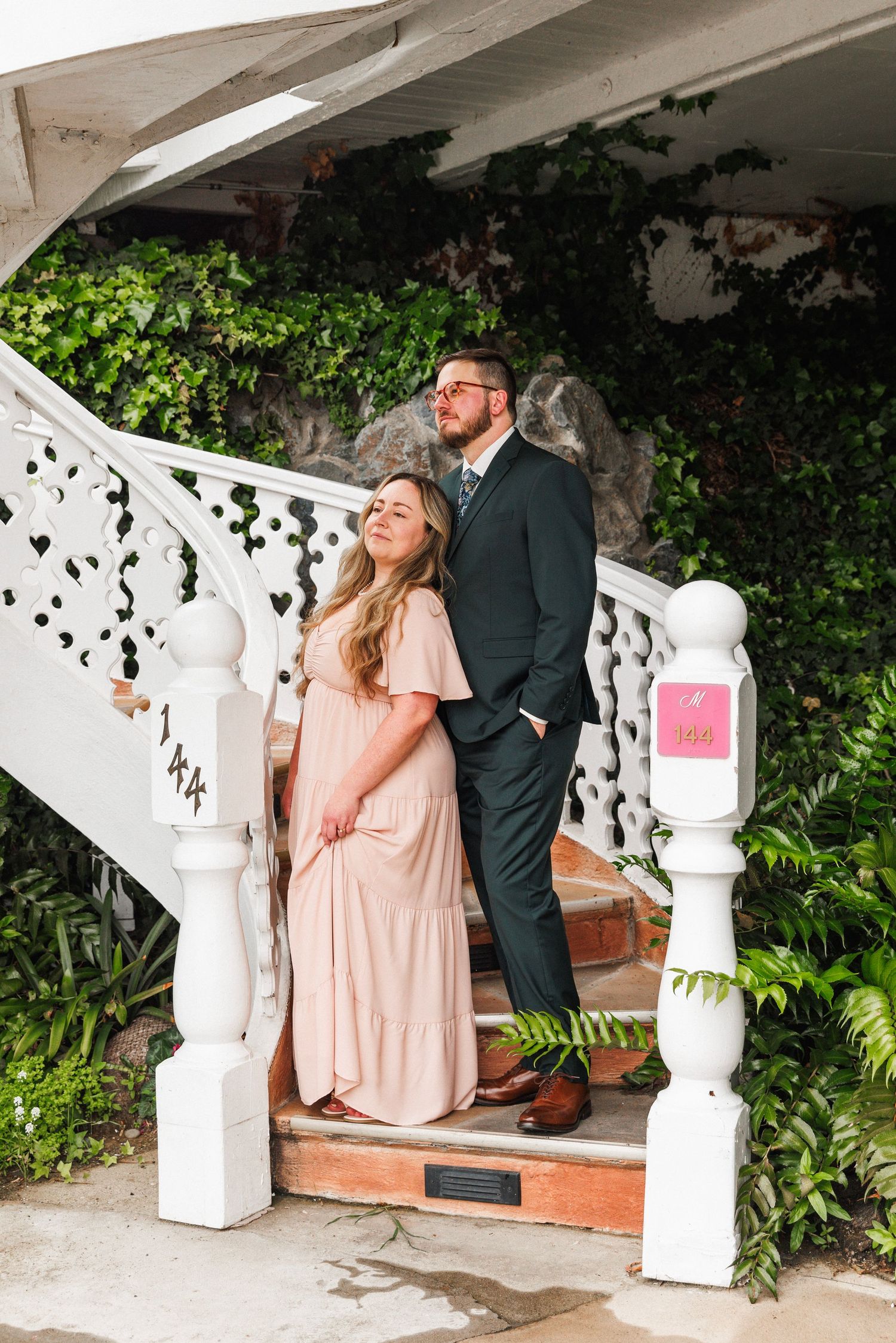 Couple poses on white ornate staircase surrounded by lush greenery, woman in pink maxi dress and man in dark suit.