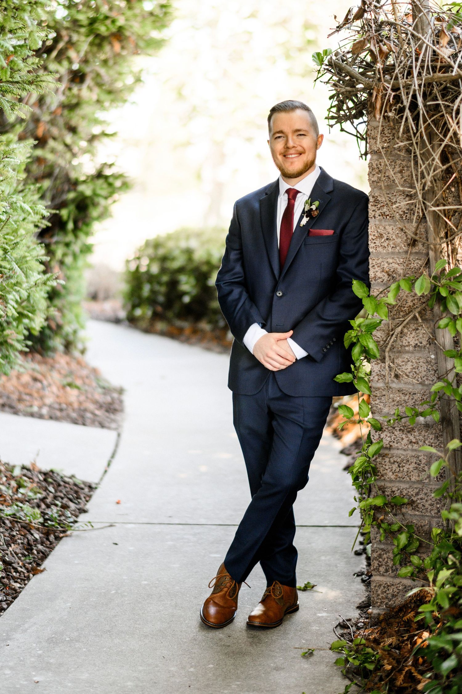 Smiling groom in navy suit with burgundy tie leaning against a tree on a garden pathway.