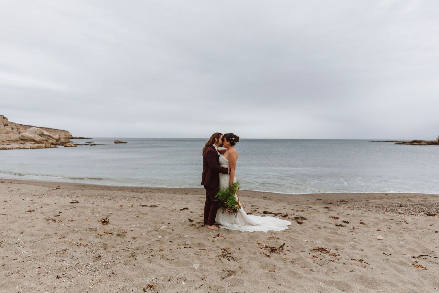 Bride and groom share a romantic kiss on a serene sandy beach with calm ocean waters behind them.
