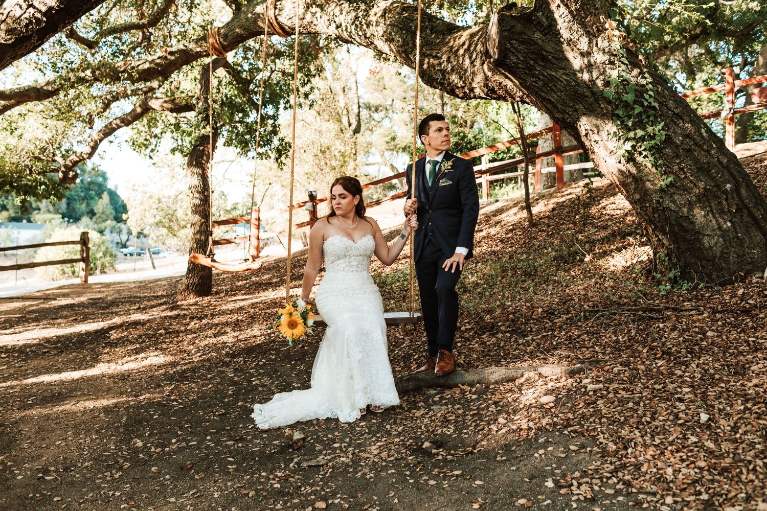 Bride and groom walk hand in hand under large oak trees on fall leaves.