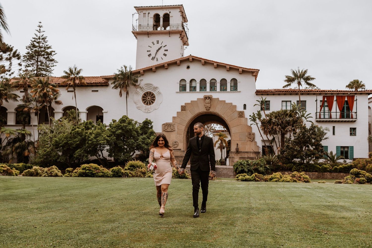 Couple walking hand in hand on a green lawn in front of the Santa Barbara County Courthouse.