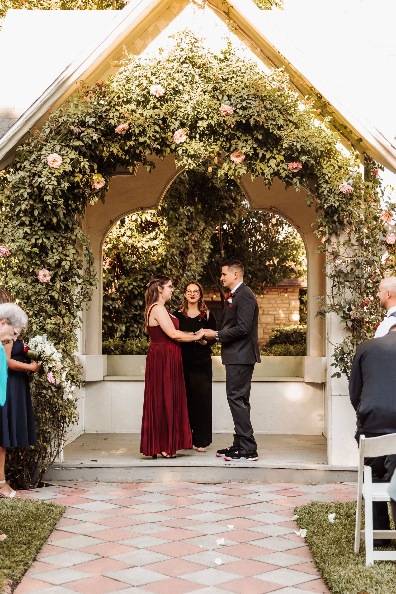 Couple exchanges vows under flower-covered arch gazebo at romantic outdoor garden wedding ceremony at dusk.
