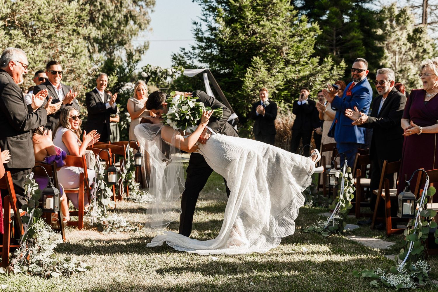 Groom dips bride joyfully at outdoor wedding ceremony while guests cheer.