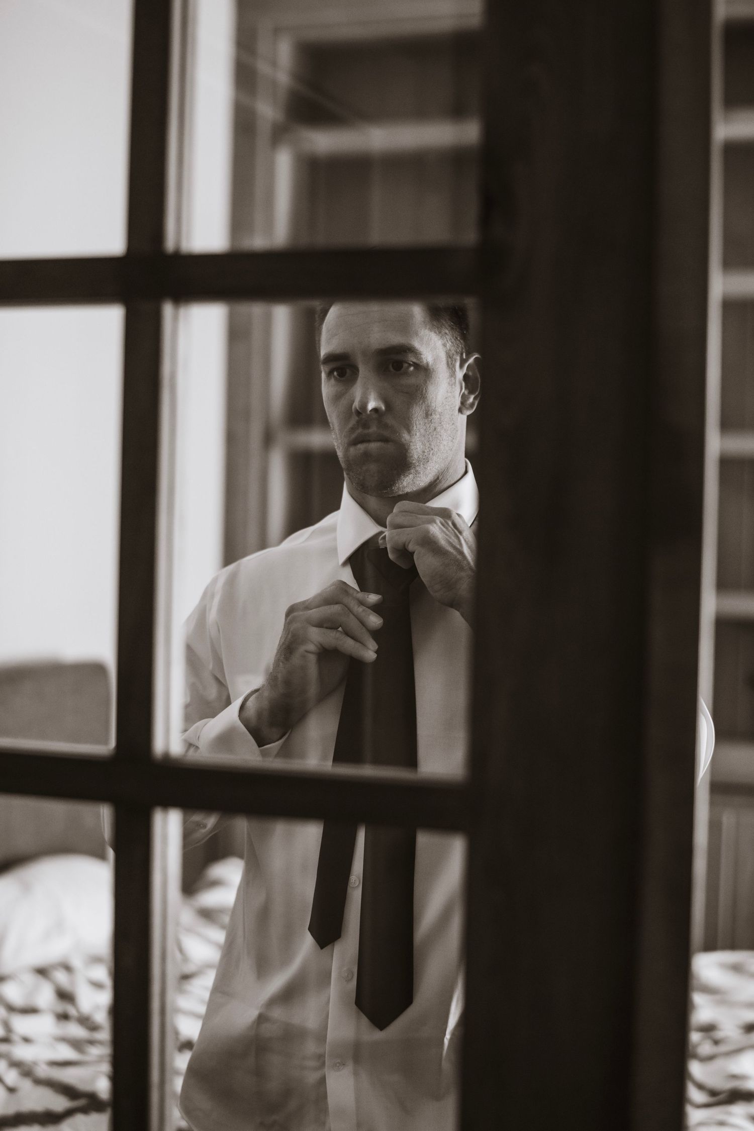 Black and white photo of a groom adjusting his tie seen through a window pane.