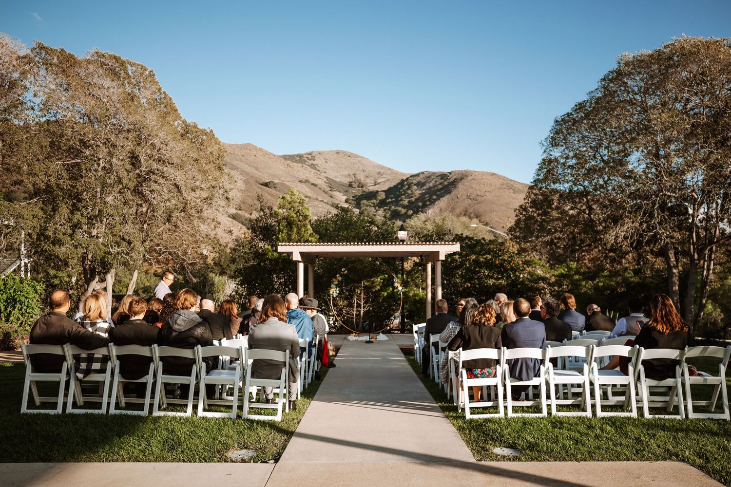 Outdoor wedding ceremony with guests seated in white chairs facing floral arch altar with mountain backdrop.