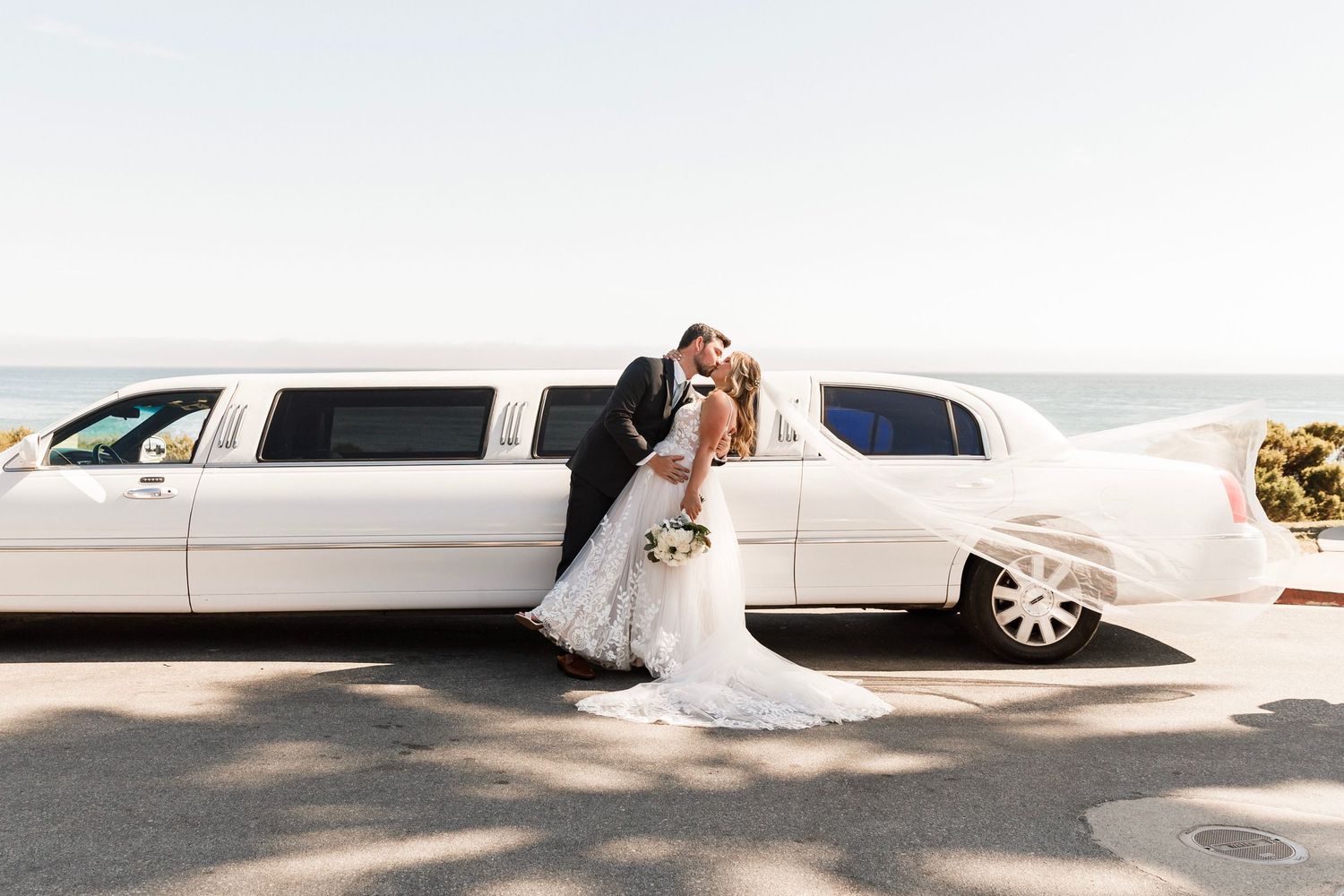 Bride and groom share a kiss leaning against white stretch limousine with ocean view backdrop.