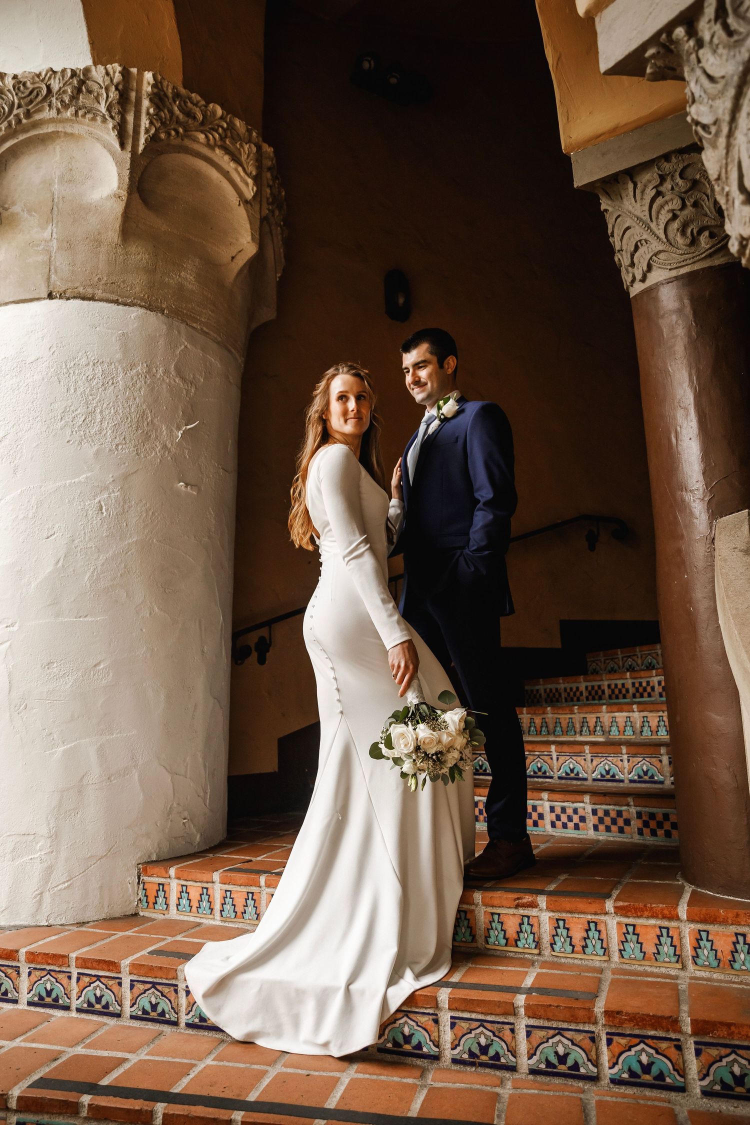 Elegant bride in a fitted white gown and groom in a navy suit posing on tiled steps with columns.